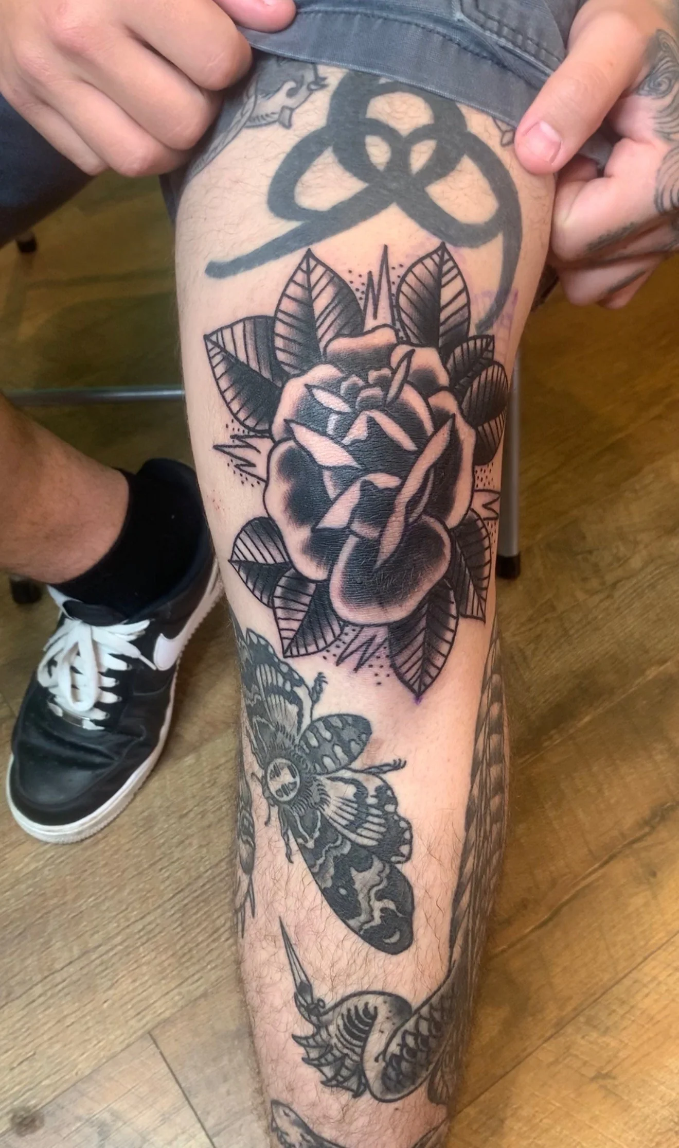 Black and grey Traditional rose tattoo with a wide leaf fan above and detailed shading, on the knee, by Tim Goodrich at his Waikiki tattoo shop, Hawaii.