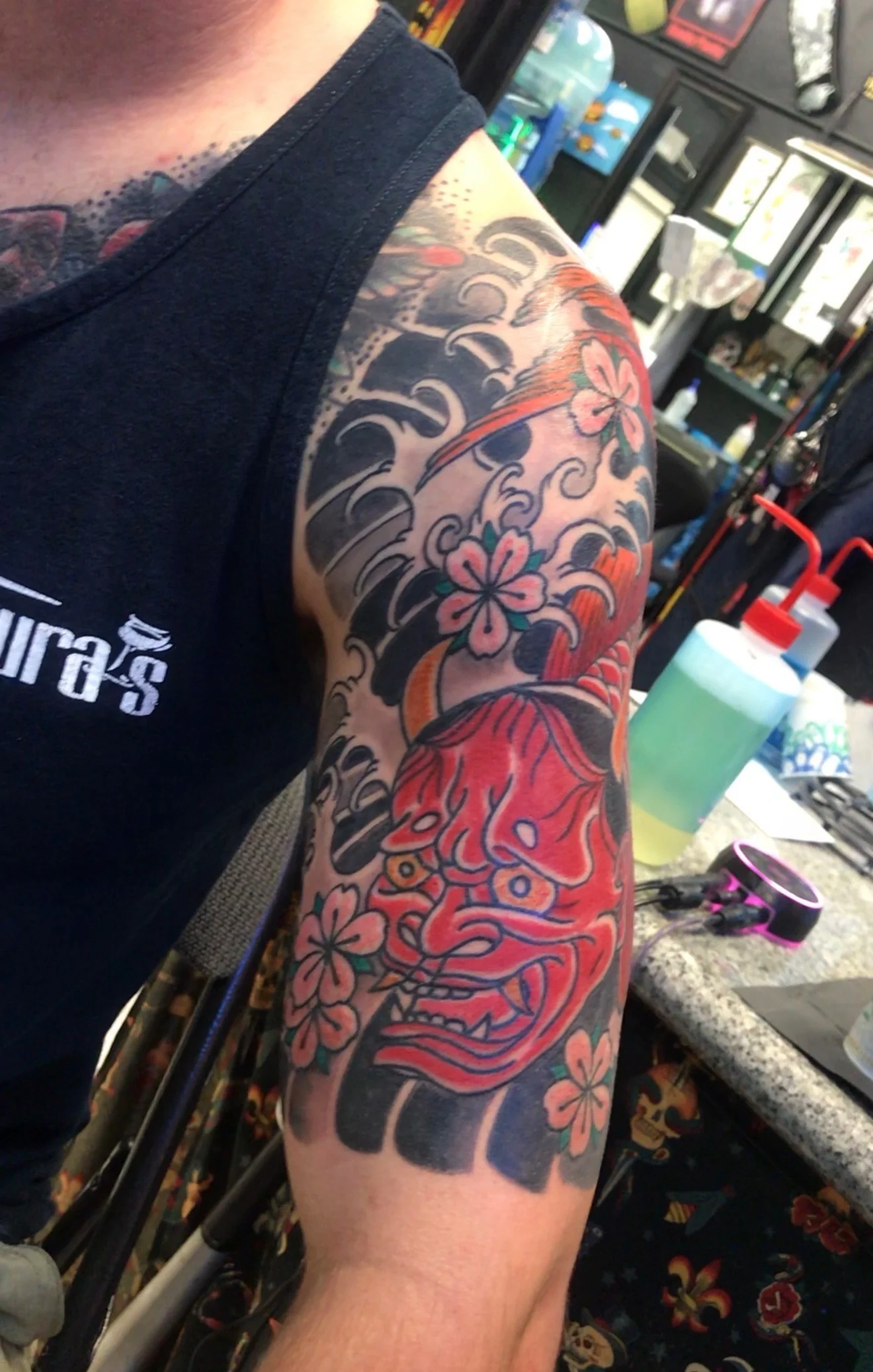 Japanese Wabori half-sleeve tattoo with a red Hannya demon mask, cherry blossoms and black wind bars on the upper arm, by Tim Goodrich at his Waikiki tattoo shop, Hawaii.