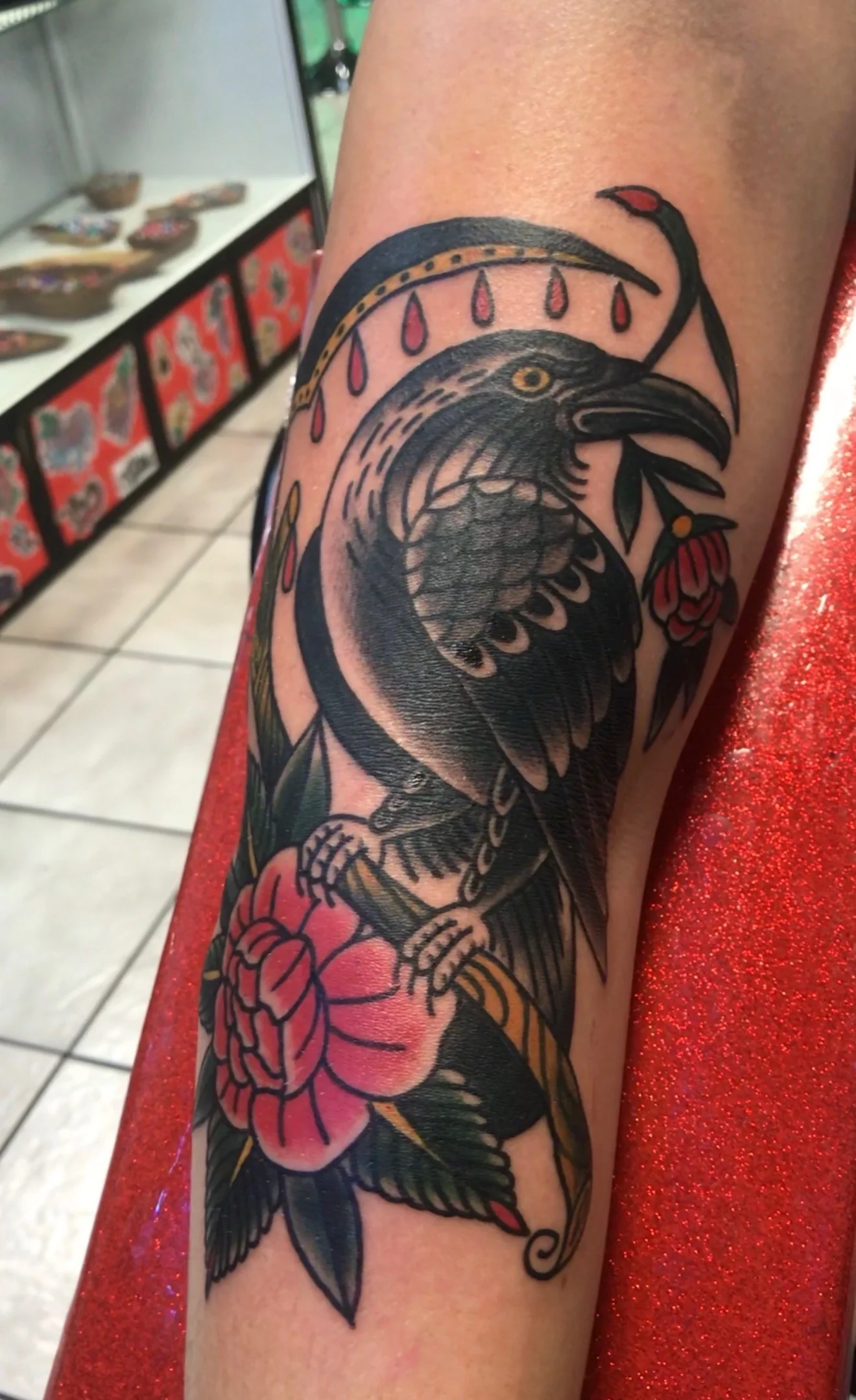 American Traditional color tattoo of a black crow perched on a thorny rose branch with a banner of dripping blood drops above, on the elbow, by Tim Goodrich at his Waikiki tattoo shop, Hawaii.